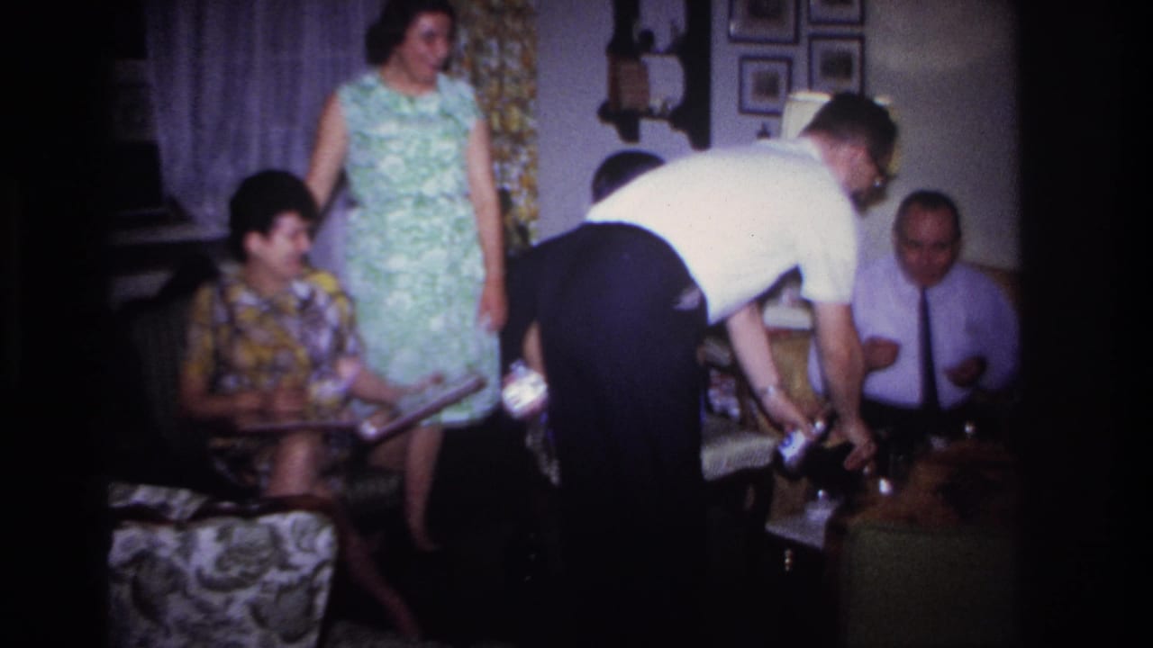 Adults gather in a living room as a man leans over a table serving drinks.