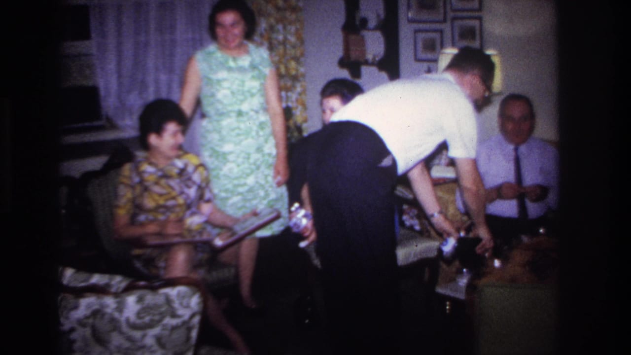 Home-movie frame from the 1967 Rockaway Beach gathering with adults around a table, including a man serving drinks in a private interior.