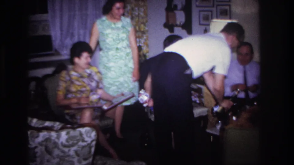 1967 domestic gathering frame showing adults in patterned 1960s clothing, seated near a low table as a man serves drinks.