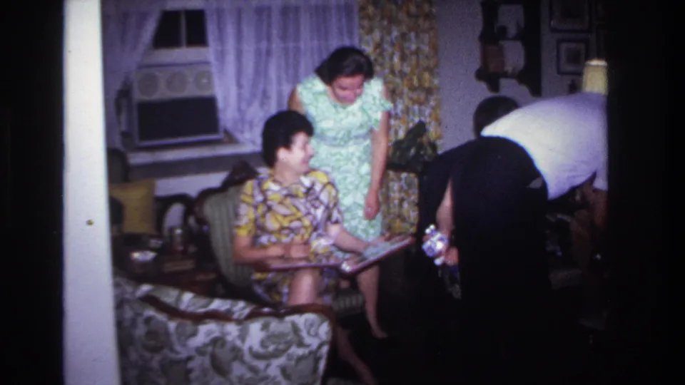 1967 Rockaway Beach living-room frame with a seated woman holding an album or folder, a standing woman, and a man serving nearby.