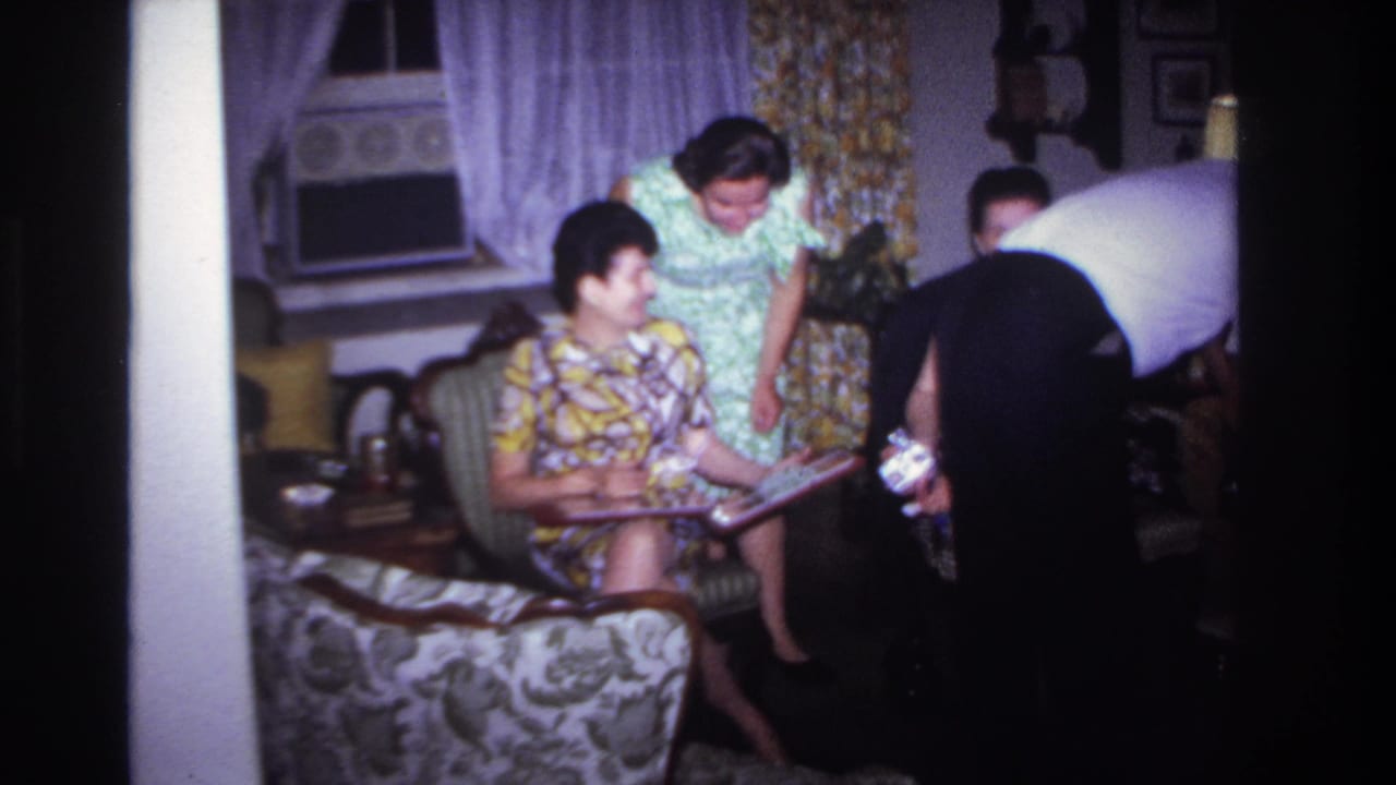 1967 Rockaway Beach home-movie frame showing adults in a living room, with a seated woman holding an album and others nearby.