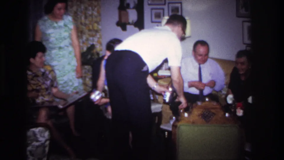 1967 Rockaway Beach home-movie frame showing relatives seated around a low table as a man serves drinks in the living room.