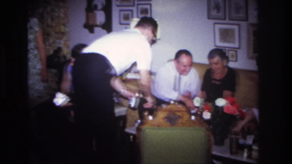 1967 Rockaway Beach home-movie frame with adults on a sofa, flowers on a table, and a man serving drinks in a private interior.