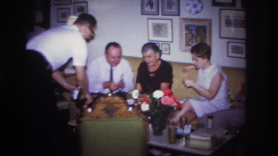 1967 Rockaway Beach home-movie frame showing adults gathered on a yellow sofa as a man serves drinks in a living room.