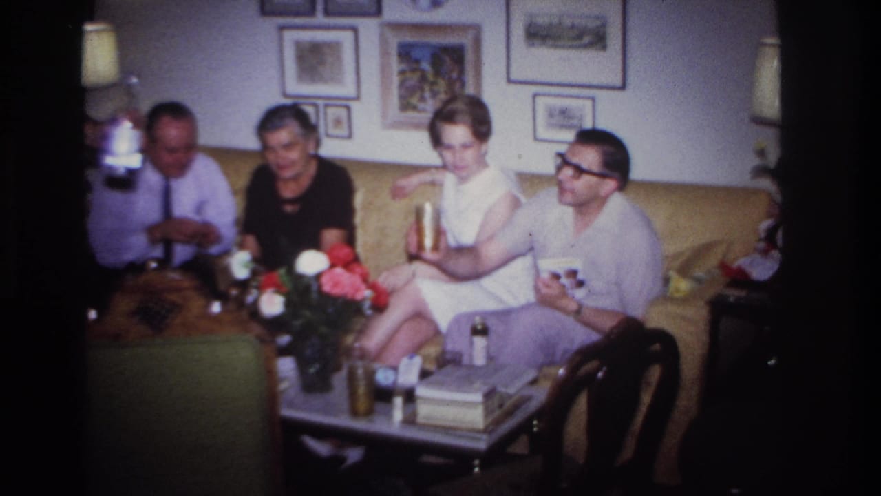 Adults in 1967 Rockaway Beach sit with drinks around a coffee table, with framed pictures and flowers visible behind them.