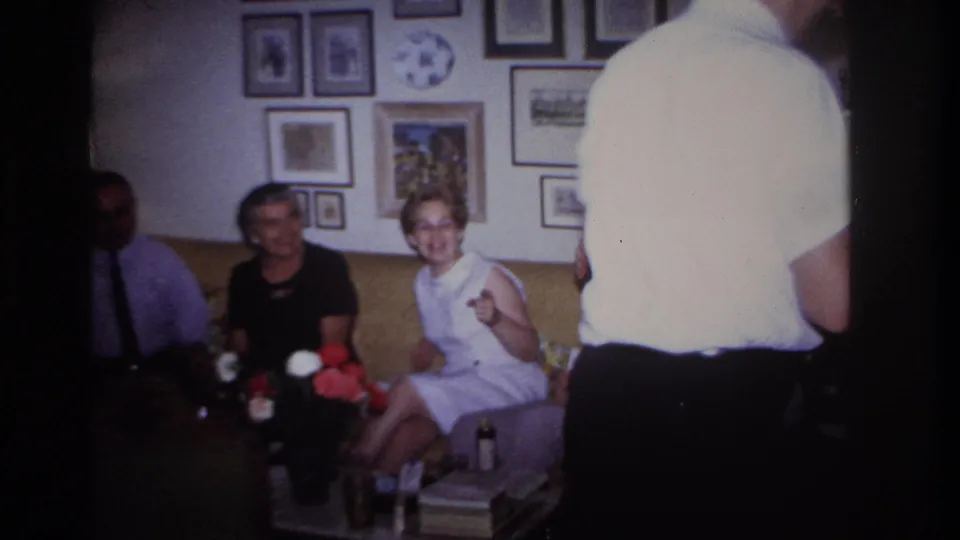 Rockaway Beach home-movie frame of smiling seated guests on a yellow sofa, partly obscured by a man standing near the camera.
