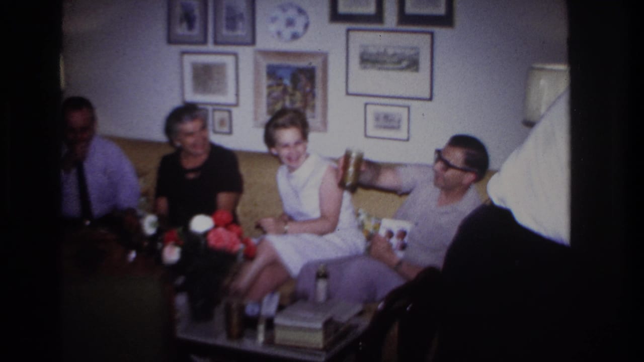 1967 Rockaway Beach home-movie frame showing four adults seated in a living room with flowers, drinks, lamps, and framed wall art.