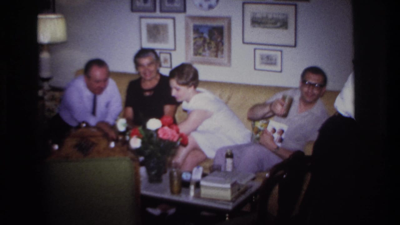 Adults sit on a couch while a man with glasses holds a drink and photo album.