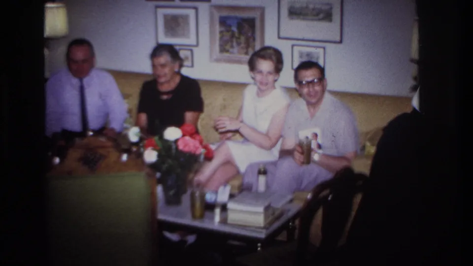 1967 Rockaway Beach home-movie frame showing four adults facing the camera in a private living room with flowers and drinks on the table.