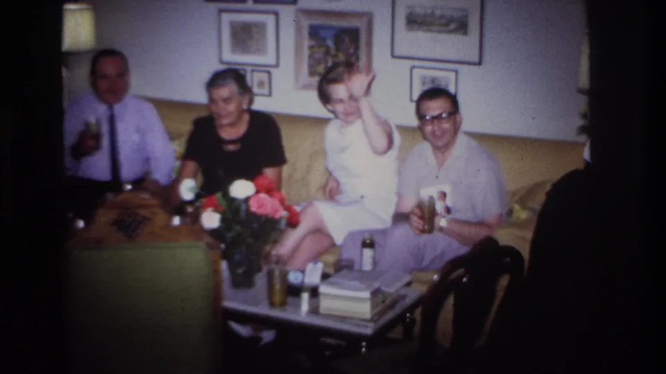 1967 Rockaway Beach frame showing a woman in a white sleeveless dress waving from a couch during a small living-room gathering.