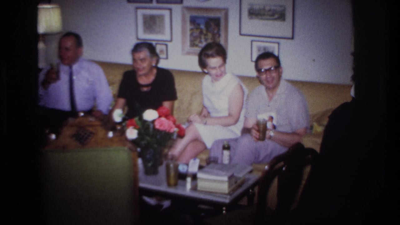 Four adults sit on a tan couch around a coffee table with flowers and bottles.