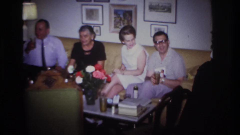1967 Rockaway Beach home-movie frame showing four adults on a tan couch in a living room with drinks, flowers, books, and framed wall art.