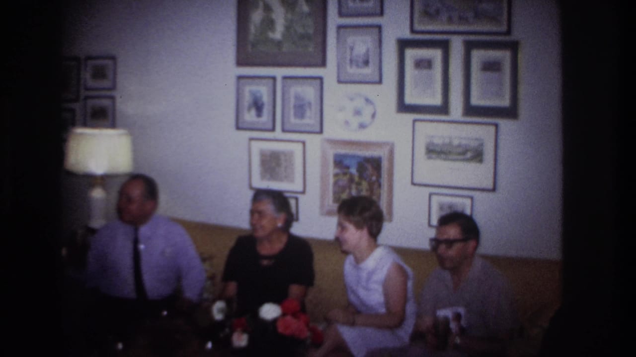 Adults in a Rockaway Beach living room sit on a tan couch, with drinks, flowers, a lamp, and framed pictures visible behind them.