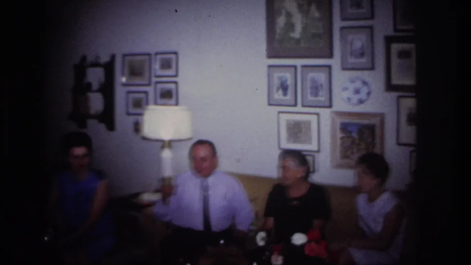 Home-movie frame from the 1967 Rockaway Beach setting showing adults seated in a living room with a lamp, framed pictures, and flowers nearby.