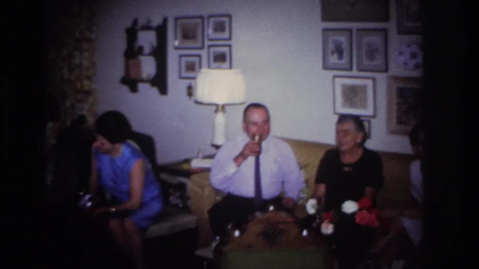 1967 Rockaway Beach home-movie frame showing a man drinking from a glass with adults seated near him, flowers and framed pictures in view.
