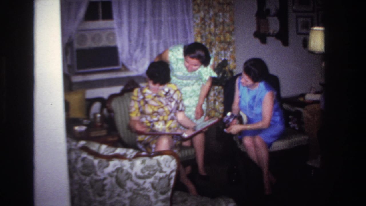 1967 Rockaway Beach home-movie frame of three women in dresses gathered around an album inside a private living room.
