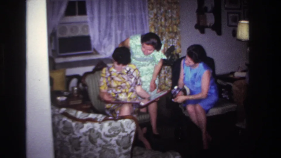 1967 Rockaway Beach home-movie frame of three women in dresses gathered around an album inside a private living room.