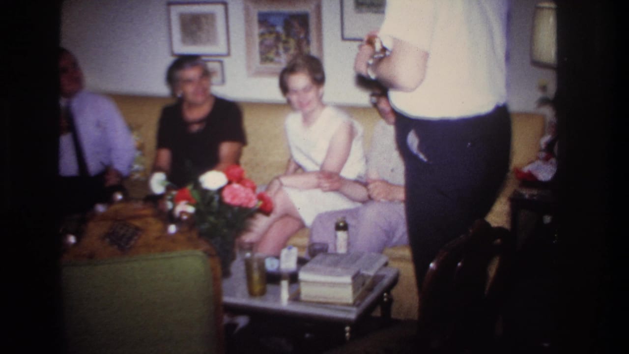 Adults sit on a yellow couch around a coffee table with flowers, glasses, and framed pictures on the wall.