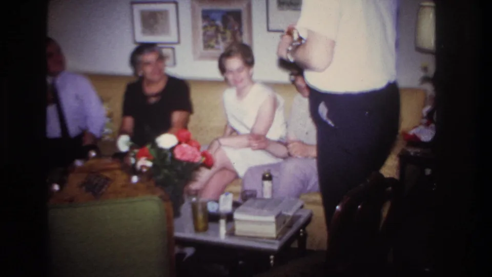 1967 Rockaway Beach living-room gathering with adults seated on a yellow couch near drinks, flowers, and framed wall art.