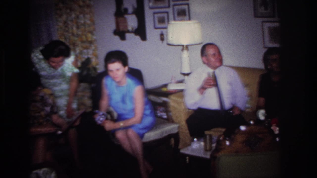 1967 Rockaway Beach home-movie frame showing adults gathered in a living room with floral curtains, lamp, sofa, and framed pictures.