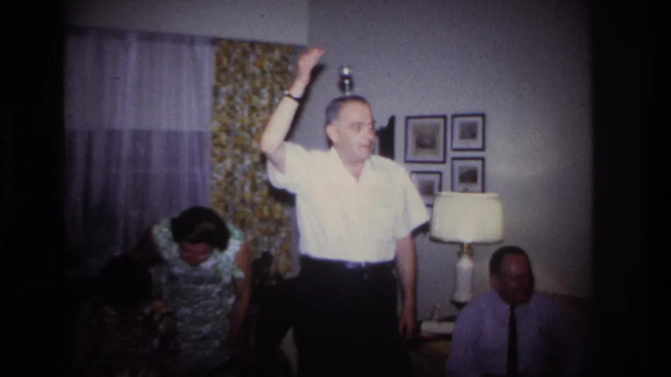 1967 home-movie frame of a man in a white shirt holding his hand above his head in a living room party scene.