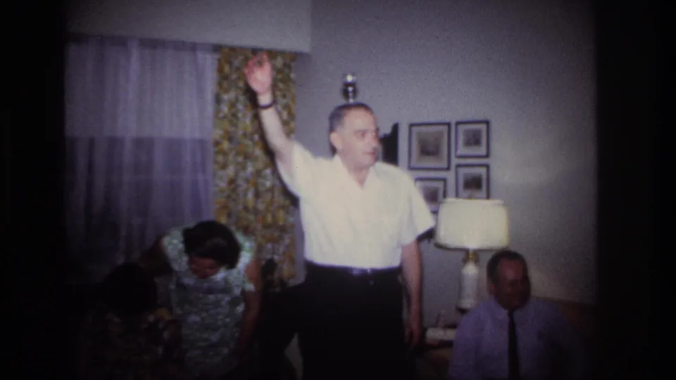 Rockaway Beach 1967 party frame showing an older man raising his arm high in a furnished living room as guests sit nearby.