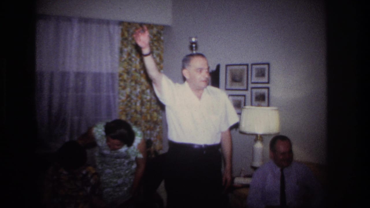 In a 1967 Rockaway Beach home-movie frame, a man in a white shirt raises one arm as adults gather around him in a private living room.