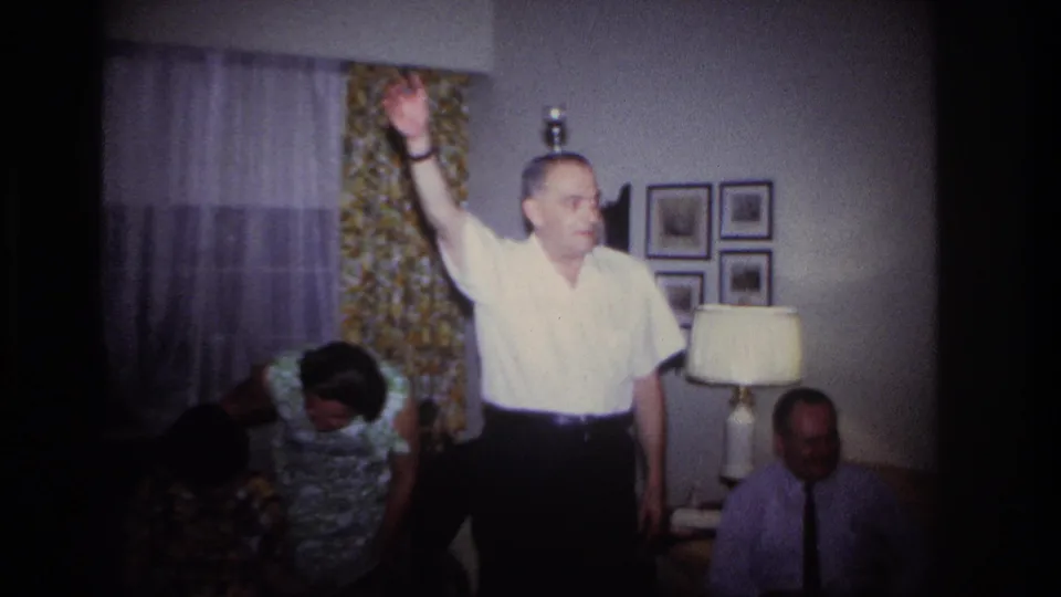 In a 1967 Rockaway Beach home-movie frame, a man in a white shirt raises one arm as adults gather around him in a private living room.