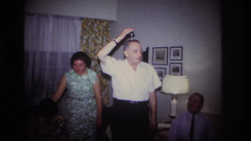 The performer appears to place a small object on his head while a woman in a green floral dress stands nearby in the Rockaway Beach living room.