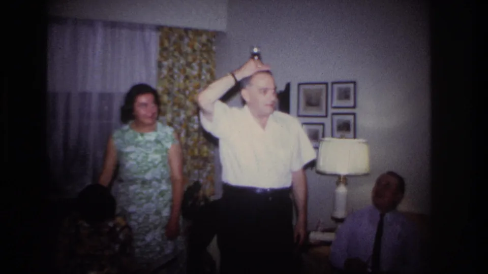 A woman in a green floral dress watches as the man continues the trick in a softly lit 1967 Rockaway Beach home interior.