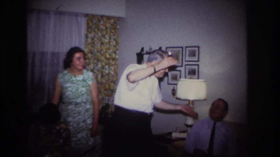 At the end of the clip, the man bends toward a seated observer while holding the small object, with lamp, curtains, and wall pictures behind them.