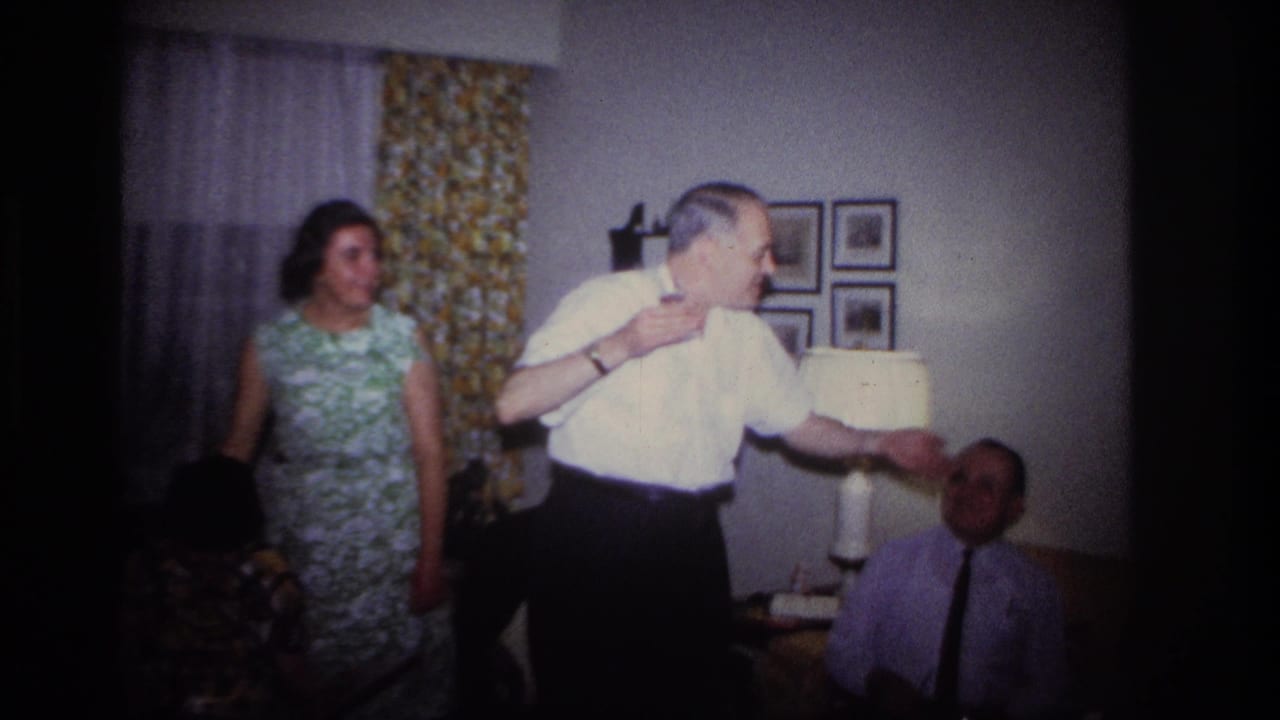 1967 Rockaway Beach home-movie frame of adults in a living room as a man gestures during a charades-like gathering.
