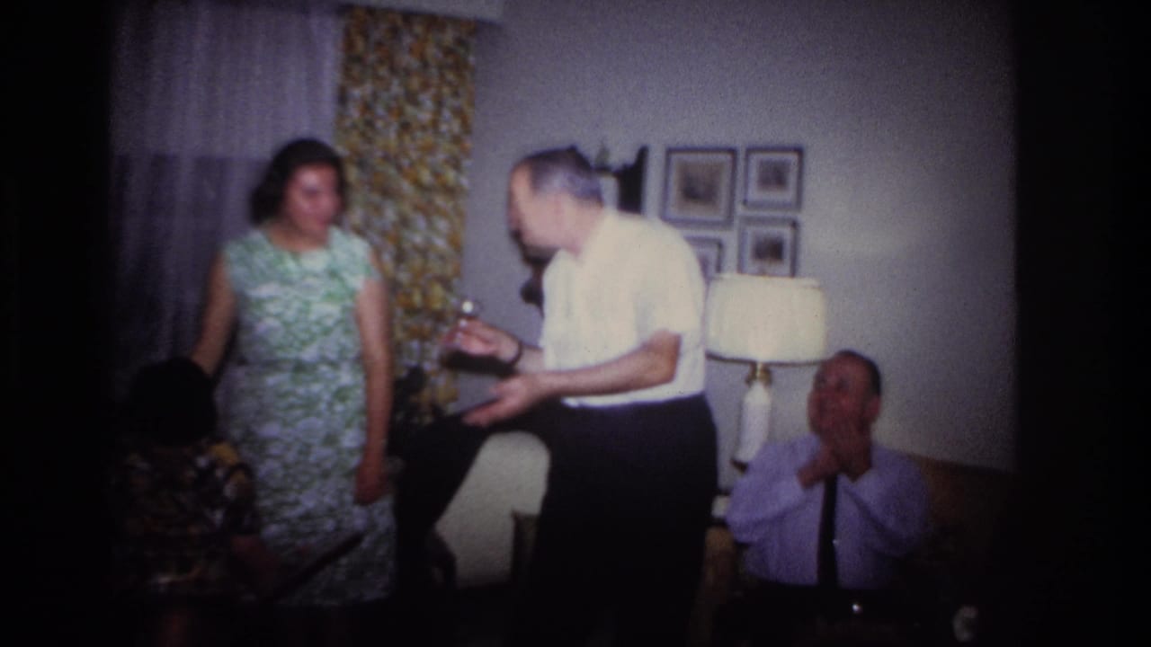 A man leans forward gesturing toward seated guests as a woman stands beside patterned curtains.