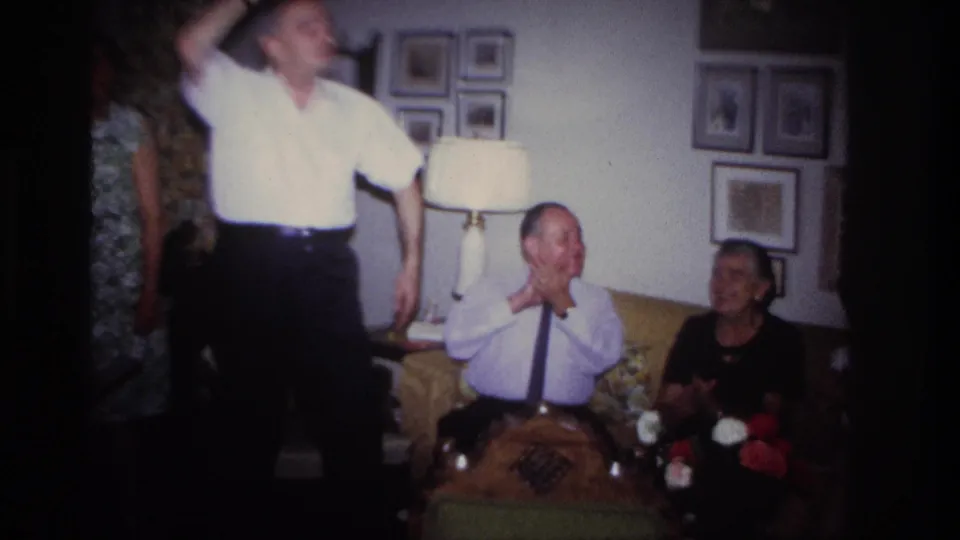 1967 Rockaway Beach home-movie frame of adults reacting as a man raises his arm during a party game.