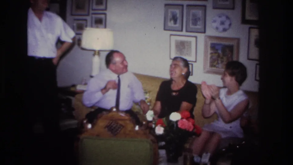 1967 Rockaway Beach frame of seated adults clapping and laughing after a charades-style moment in a private living room.
