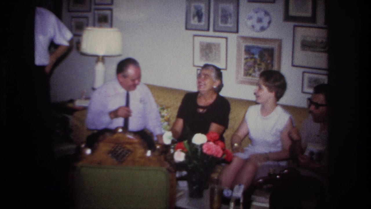1967 Rockaway Beach home-movie frame of adults gathered on a sofa in a living room with flowers, lamp, framed pictures, and a decorative plate.