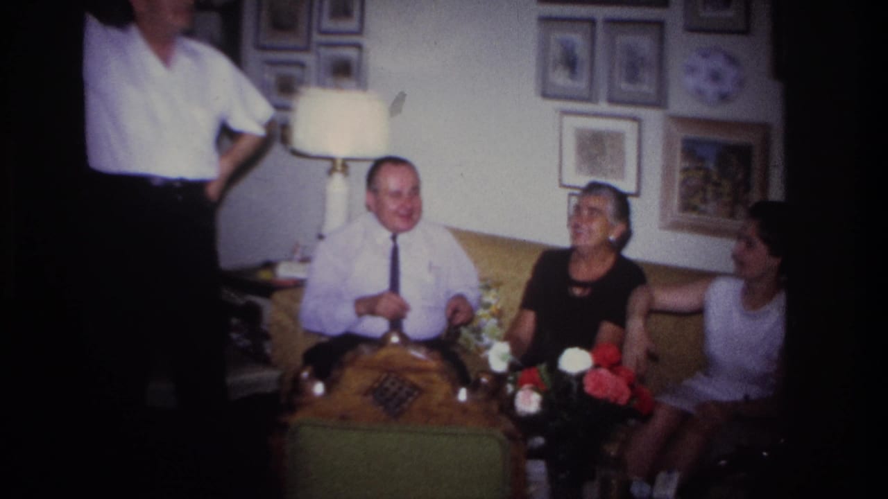Seated adults smile and gesture on a sofa, with a lamp, flowers, and framed pictures behind them.
