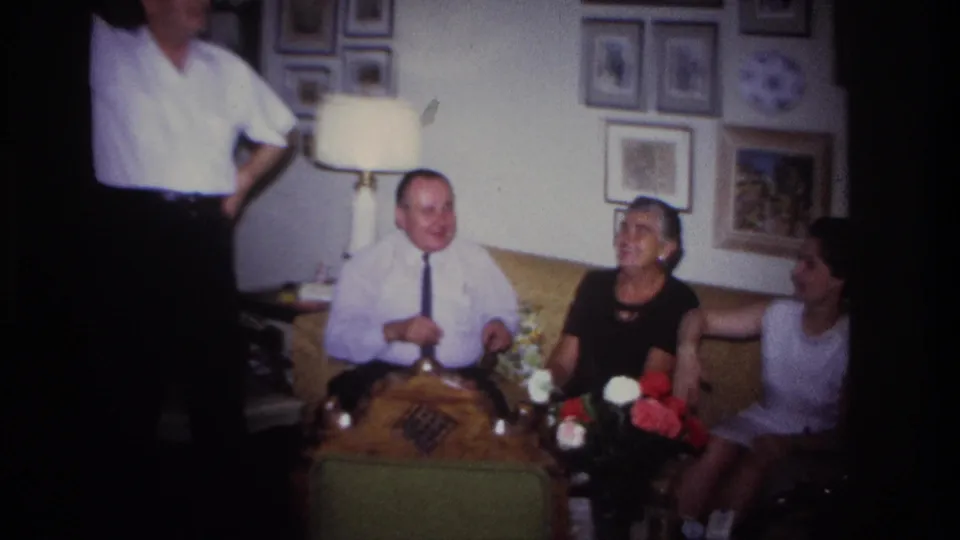 Adults in 1967 Rockaway Beach appear to laugh and converse on a sofa in a private living room with a lamp, flowers, and framed pictures.