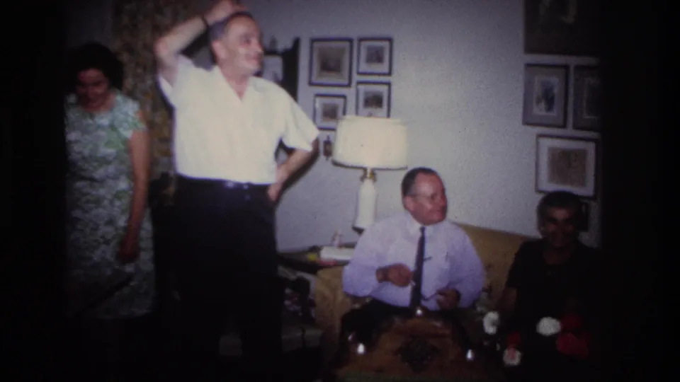 In a 1967 Rockaway Beach living-room scene, a man stands with one hand raised while others sit nearby and a woman in a patterned dress stands at left.