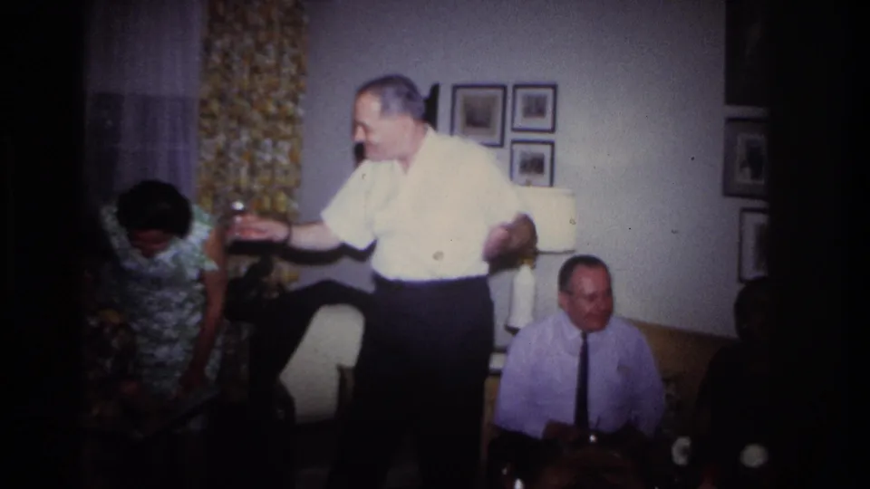 A 1967 Rockaway Beach home-movie frame shows a man gesturing animatedly in a living room as seated and standing adults look on.
