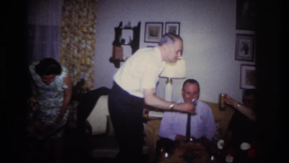 Rockaway Beach 1967 home-movie frame showing adults in a living room during a toast, with framed pictures and patterned curtains behind them.