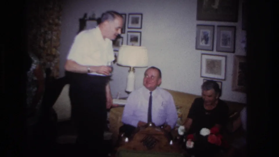 Rockaway Beach 1967 home-movie still with a standing man holding a glass beside seated adults, flowers, and framed wall decor.