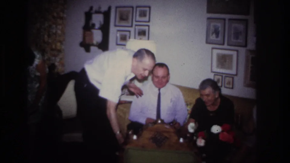 Rockaway Beach 1967 frame showing a man leaning over a seated guest during a living-room gathering with flowers and wall photographs visible.