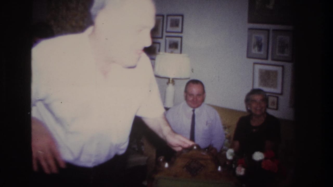 The performer moves close to the camera, partly overexposed, with two seated relatives visible behind him in the living room.