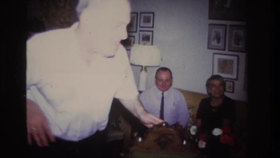 The performer moves close to the camera, partly overexposed, with two seated relatives visible behind him in the living room.
