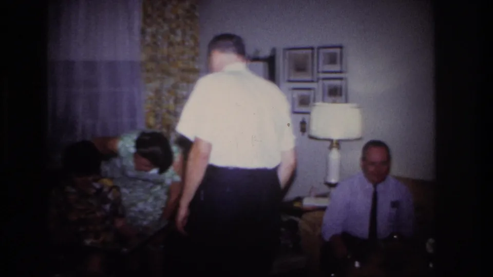 The man stands with his back to the camera in the living room, with other adults seated nearby and wall photos behind them.
