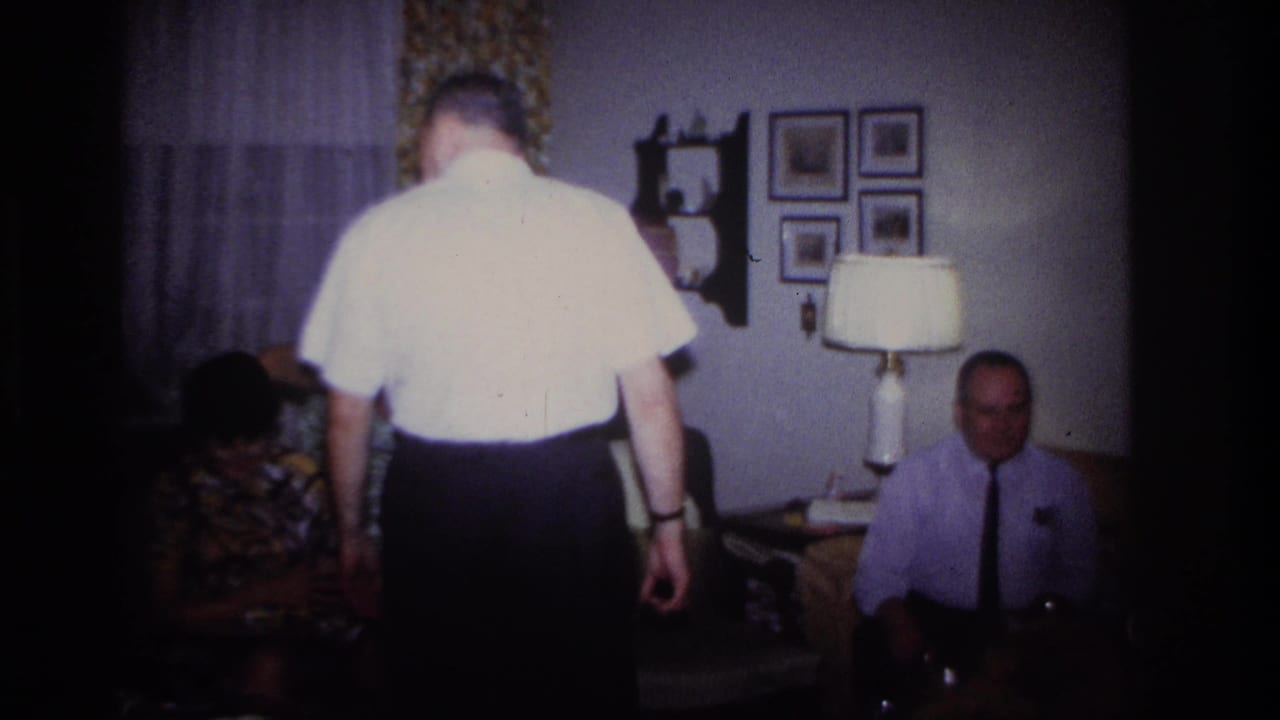 1967 Rockaway Beach home-movie frame showing adults in a living room with a lamp, framed pictures, curtains, and floral seating.