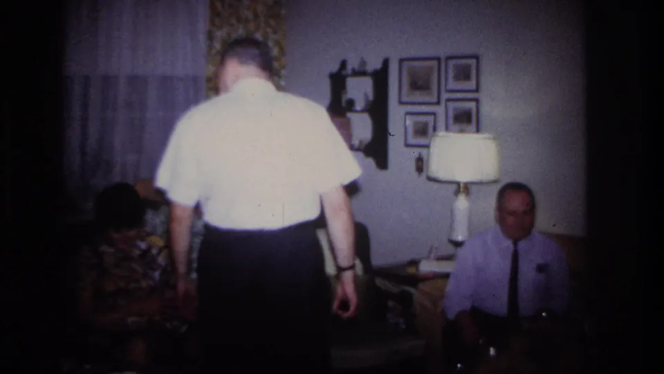 1967 Rockaway Beach home-movie frame showing adults in a living room with a lamp, framed pictures, curtains, and floral seating.