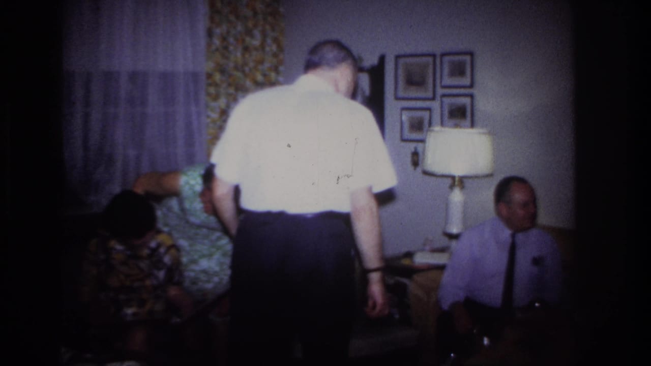 Adults gather in a living room as a man stands facing away and others sit near a lamp and framed pictures.