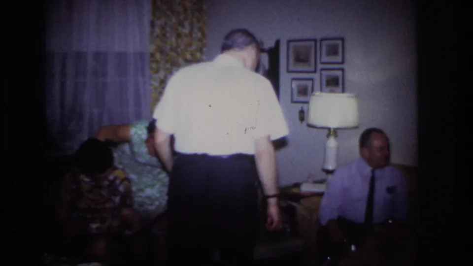Rockaway Beach 1967 frame with adults in a private living room, including a standing man, seated figures, lamp, curtains, and wall pictures.